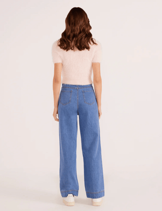 A person with wavy brown hair wears the MINKPINK Millie Fluffy Knit Tee in light pink, paired with blue high-waisted wide-leg jeans and white sneakers, standing with their back to a plain light background.