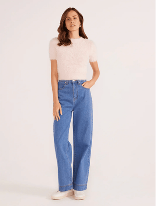 A woman stands against a light background, wearing the MINKPINK Millie Fluffy Knit Tee in pale pink with high-waisted blue jeans and white shoes. One hand is in her pocket as she glances slightly to the side.