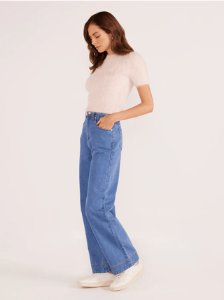 A woman stands sideways, looking down, wearing the MINKPINK Millie Fluffy Knit Tee in light pink with high-waisted blue jeans and white sneakers. This cozy top is a perfect transitional piece for any season.