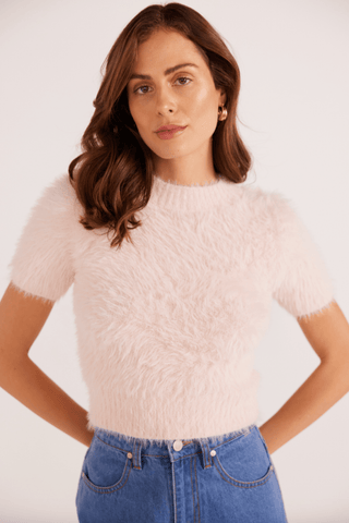 A woman with wavy brown hair models the MINKPINK Millie Fluffy Knit Tee in pale pink, paired with high-waisted blue jeans. She stands against a plain light background, arms slightly behind her, looking at the camera.
