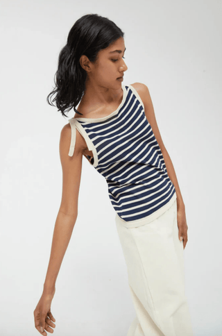 A woman with medium-length dark hair poses against a light background wearing the Compania Fantastica Nautical Stripe Knit Tank Top-FINAL SALE with cream trim and high-waisted white pants, her head tilted and one arm extended.