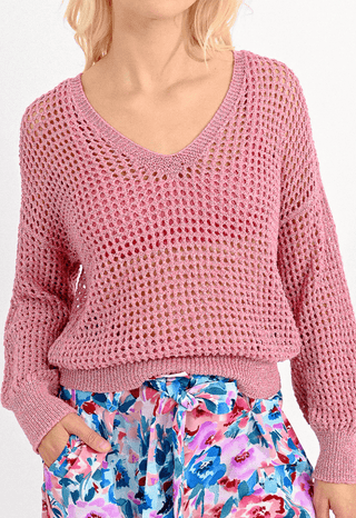 A person with light blonde hair wears the Molly Bracken Open Weave V-neck Knit Sweater in light pink, styled with high-waisted blue, pink, and purple floral pants and has one hand in their pocket.