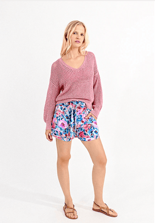 A woman with blonde hair poses against a white background, wearing the Molly Bracken Open Weave V-neck Knit Sweater in pink, paired with colorful floral shorts and brown sandals. She has a relaxed expression and one hand in her pocket.