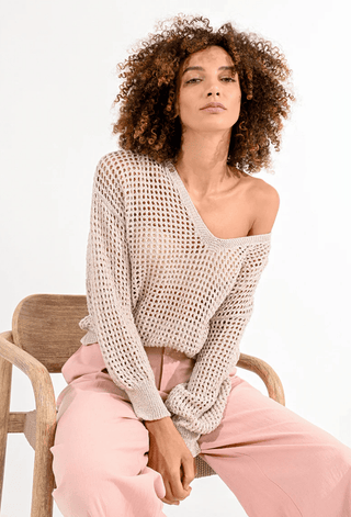 A woman with curly hair sits on a wooden chair, wearing the Molly Bracken Open Weave V-neck Knit Sweater in beige and light pink pants, styled off one shoulder, set against a plain white background.