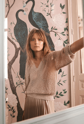 A young woman with light brown hair wears the Molly Bracken Open Weave V-neck Knit Sweater-FINAL SALE and a pleated skirt, standing indoors before a wall adorned with two large peacocks and white flowers.