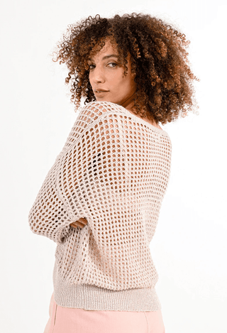 A person with curly hair wears the Molly Bracken Open Weave V-neck Knit Sweater–FINAL SALE over pink pants, standing against a plain white background and glancing over their shoulder.