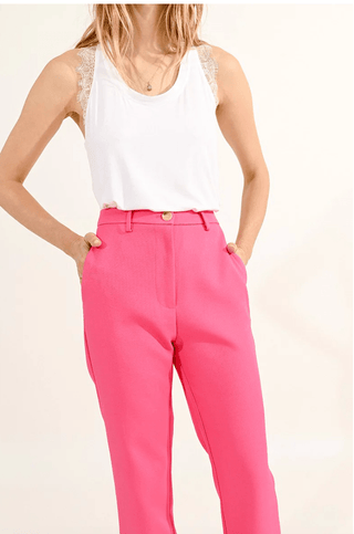 A woman stands against a plain light background wearing the Molly Bracken Priscilla Pants in pink and a sleeveless white lace-detailed top, with her hands in the pockets.