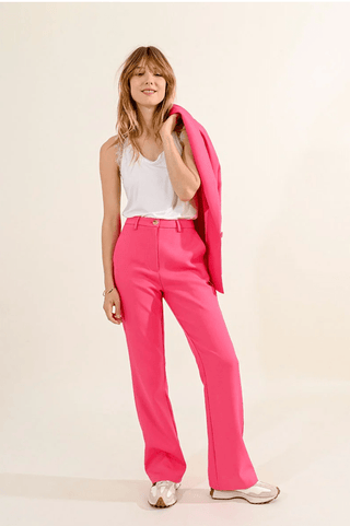 A woman smiles in a white sleeveless top, Molly Bracken Priscilla Pants-FINAL SALE in bright pink, and white sneakers, holding a matching pink blazer over her shoulder against a plain light background.