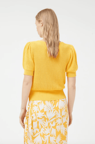 A woman with blonde hair, seen from behind, wears the Compania Fantastica Short Sleeve Knit Sweater-FINAL SALE in yellow and a white skirt with a yellow botanical print, set against a plain light background.