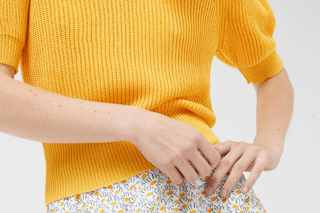 A person wears the Compania Fantastica Short Sleeve Knit Sweater-FINAL SALE in yellow with a white skirt featuring a yellow and gray daisy pattern, adjusting the skirt. The image highlights their torso and hands.