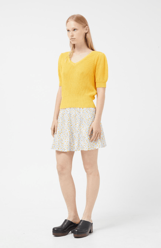 A woman with straight light brown hair and a neutral expression wears the Compania Fantastica Short Sleeve Knit Sweater-FINAL SALE in bright yellow, paired with a white floral skirt and black platform shoes against a plain white background.