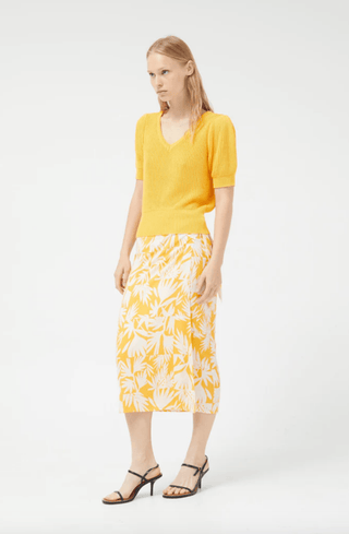 A person in a bright yellow Compania Fantastica Short Sleeve Knit Sweater-FINAL SALE, a yellow and white floral midi skirt, and black strappy heels stands against a plain white background.
