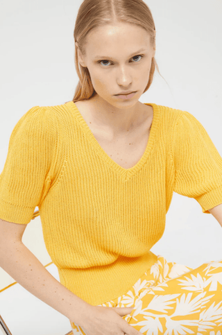 A woman with straight blonde hair wears Compania Fantasticas Short Sleeve Knit Sweater-FINAL SALE in yellow and a white skirt with a bold yellow leaf pattern, sitting on a light chair against a plain white background.