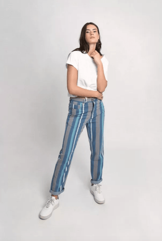 A woman with long, dark hair poses confidently in Molly Bracken Stripe Denim Pants-FINAL SALE (blue and gray), a white T-shirt, and white sneakers against a plain light background.