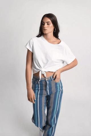 A woman with long dark hair wears a white knotted t-shirt and Molly Brackens Stripe Denim Pants-FINAL SALE in blue-and-teal vertical stripes, standing against a plain light background.