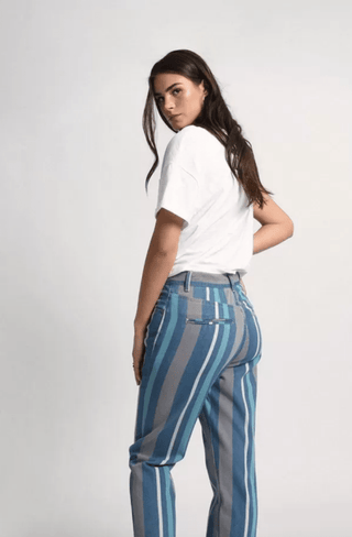 A woman stands sideways, looking over her shoulder in a white t-shirt and Molly Bracken Stripe Denim Pants-FINAL SALE with vertical blue and gray stripes, set against a plain light background.