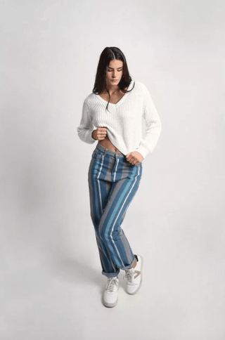 A woman poses against a plain backdrop, wearing a white knit sweater, white sneakers, and Molly Brackens Stripe Denim Pants-FINAL SALE. She looks down and holds the hem of her sweater with one hand.
