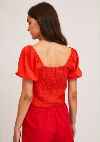 A woman with long brown hair is seen from the back, wearing Compania Fantasticas Smocking Crop Top With Puff Sleeves-STREET SALE in bright orange, paired with matching shorts against a plain light background.