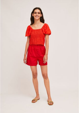 A woman smiles against a plain background, wearing the Compania Fantastica Smocking Crop Top With Puff Sleeves-STREET SALE in orange, paired with red shorts and gold flat sandals.