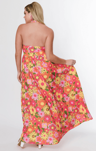 A woman with long blonde hair models the Smak Parlour Flower Power Keyhole Halter Maxi Dress, a vintage-inspired, brightly floral floor-length style. Facing away, she holds part of the skirt against a plain white background.