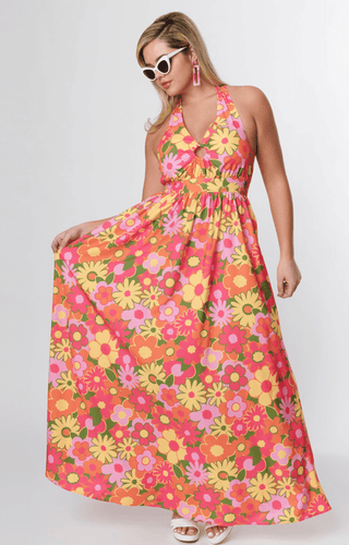 A woman models the Smak Parlour Flower Power Keyhole Halter Maxi Dress-FINAL SALE, featuring a bold pink, yellow, orange, and green floral print. She accessorizes with white sunglasses, pink earrings, and white sandals.