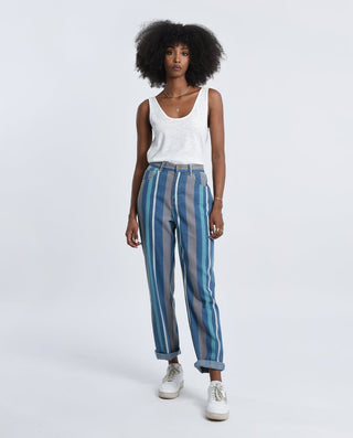 A woman with curly hair models Molly Bracken’s Stripe Denim Pants-FINAL SALE—blue and gray vertical stripes—paired with a white sleeveless top and sneakers, set against a plain light gray background.