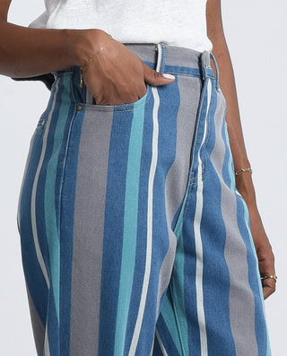 A person models Molly Bracken’s Stripe Denim Pants-FINAL SALE, featuring vertical blue, teal, and gray stripes, paired with a white top and delicate gold bracelets, standing with one hand in a pocket.
